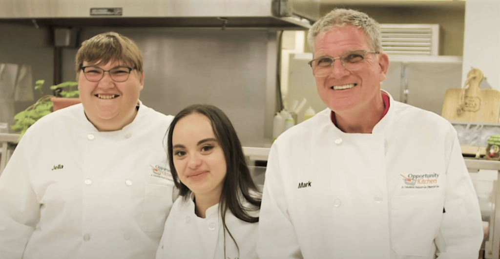 Video thumbnail showing three smiling people wearing chef's coats with the Opportunity Kitchen logo while standing in an industrial kitchen prep area.